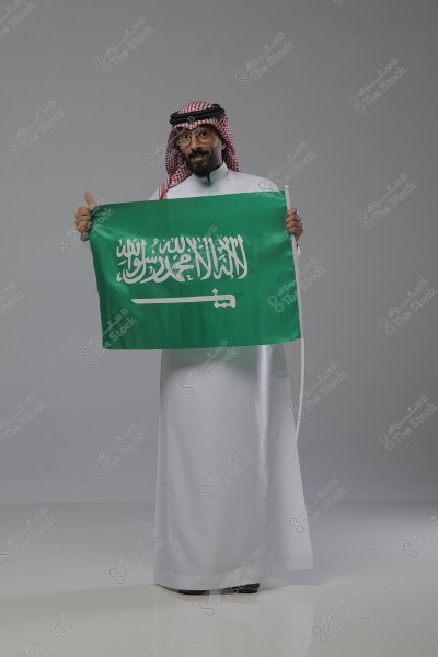 An image of a man wearing traditional Saudi clothing, consisting of a white thobe, red and white checkered ghutra, and a black agal. He is holding the green flag of Saudi Arabia, which features the Shahada and a white sword. The man is standing in a studio with a simple gray background.