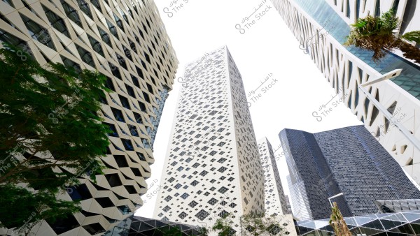 An image of a group of modern buildings with unique architectural designs. The building in the foreground features a white facade with a complex pattern of triangles and grids. Surrounding buildings have glass facades and various geometric patterns. Green trees are seen at the bottom, adding contrast to the scene.