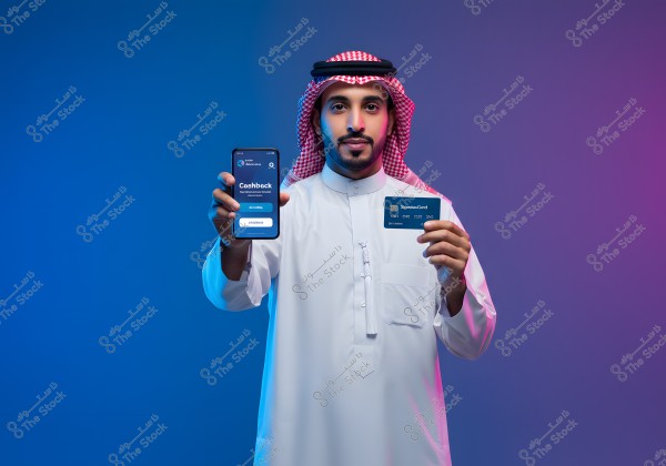 An image of a man wearing traditional Saudi attire, featuring a red and white headscarf and a white robe. He is standing against a blue and purple gradient background, holding a mobile phone displaying an app with the word \"Cashback\" and a blue credit card in the other hand.
