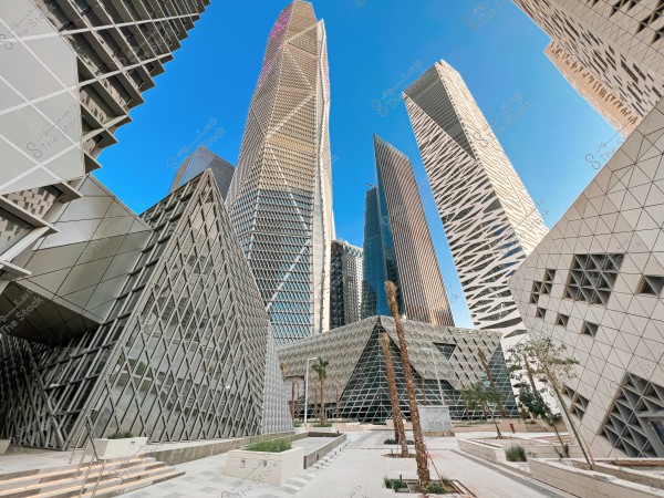 View of several modern and tall buildings in a city, featuring unique architectural designs with sharp angles and glass and metal facades. The background shows a clear blue sky, and palm trees adorn the open spaces between the buildings.