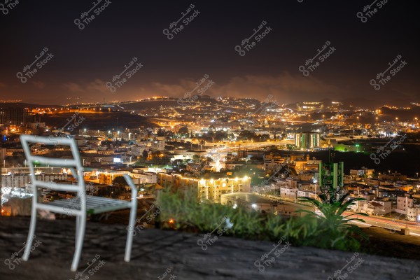A nighttime cityscape with a brightly lit city in the background. In the foreground, there is an empty metal chair next to a patch of greenery. The city lights illuminate buildings and winding streets, with dark hills visible in the distance.