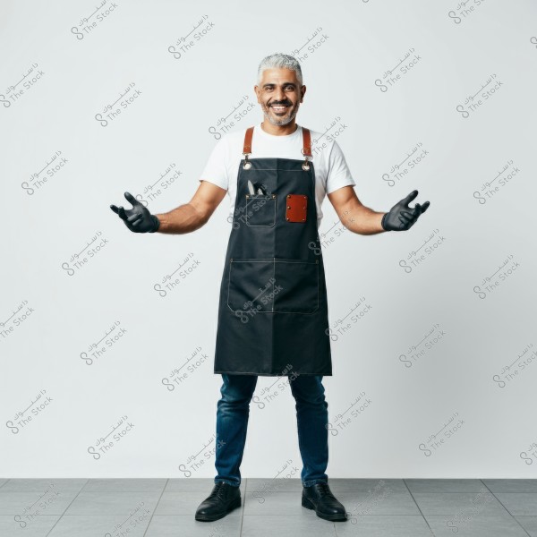 A smiling man with gray hair wearing a white shirt and a black apron with pockets and brown straps, standing against a white background. He is wearing black gloves on his hands, blue jeans, and black shoes.