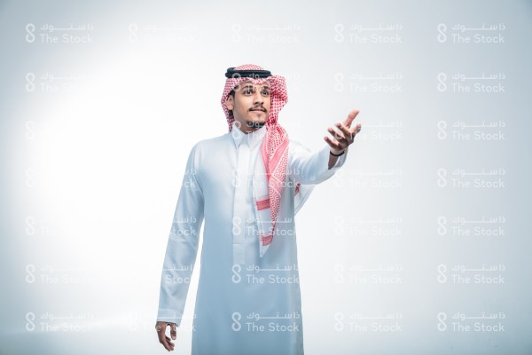 بورتريه رجل سعودي يرتدي الثوب ويقف أمام خلفية بيضاء، يتفاعل بإشارات تعبيرية بيدية ، يمد يده ليمسك بأحدهم ، يمد يد المساعده ، مساعدة الآخرين ، صورة شخصية