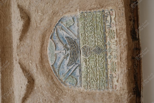 Image of a wall featuring an ornate decorative panel belonging to Islamic art, carefully carved into clay or plaster. The panel displays a semi-circular design with Arabic scripts, botanical motifs, and geometric curves in shades of green, blue, and beige. The wall appears aged with a natural, rough texture.