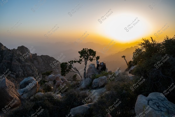 An Image of  mountains at sunset, featuring green plants and large rocks in the foreground. In the middle, a group of people are sitting on the rocks, watching the sunset. The sun is setting over the mountains in the background, casting shades of orange and blue across the sky. This image is taken from Al Shafa mountains, Taif Reagions, Saudi Arabia
