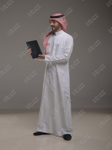يظهر في الصورة رجل يرتدي ثوبًا أبيض وغترة حمراء مع عقال, وينظر إلى جهاز لوحي يحمله بيديه. الخلفية ذات لون رمادي محايد. يُعتقد أن الملابس من الطراز السعودي التقليدي.