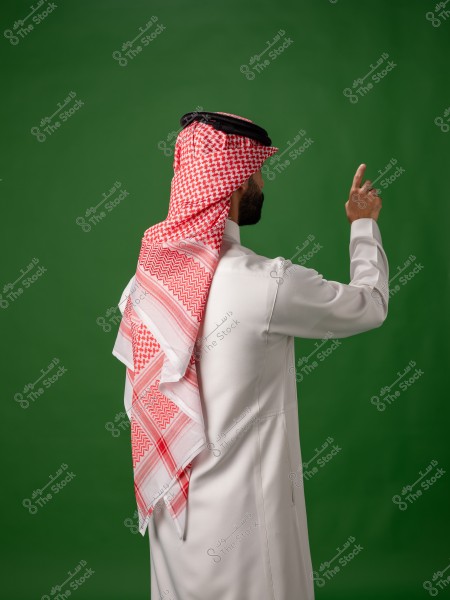 A man wearing traditional Saudi thobe and ghutra stands in front of a green background, pointing with his hand.