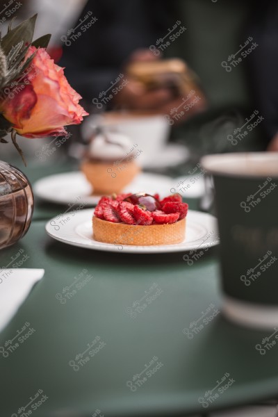 A tart filled with red raspberries served on a white plate with a blurred background featuring a cup, other dishes, and a flower in a vase.