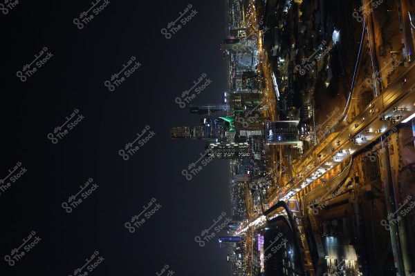 A nighttime image of Riyadh, Saudi Arabia, showing the city skyline illuminated with lights from tall buildings and busy streets filled with cars. The Kingdom Tower is clearly visible with lights surrounding it, along with various roads and bridges lit up at night. The dark sky accentuates the bright city lights.