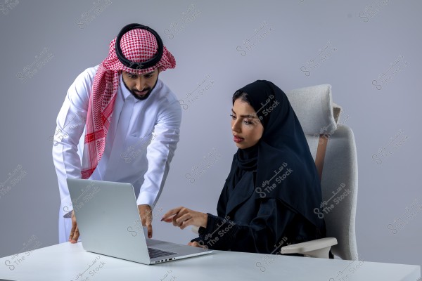 A man wearing traditional Saudi attire, including a thobe, ghutra, and agal, stands next to a woman in a black abaya and hijab, both looking at a laptop screen. They are in an office environment with a gray background. The woman is seated in a comfortable chair, working on the laptop, while the man points at the screen.