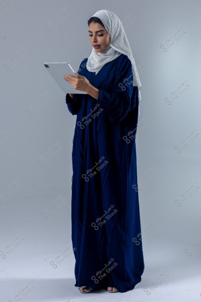 A portrait of a woman wearing a blue abaya and a white hijab, standing in a studio with a light gray background, focusing on using an electronic tablet with her hands.