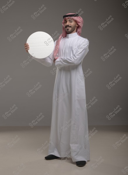 Portrait of a man wearing traditional white Arabic thobe and a red and white checkered ghutra with a black agal. The man stands in a relaxed pose, holding a circular board in his upper hand. The background is grey.