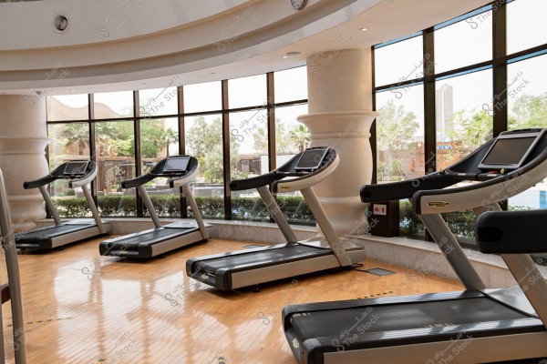 Image of an indoor gym equipped with modern treadmills arranged neatly on a shiny wooden floor. The walls are large glass windows offering a view of outdoor scenery with trees and plants. The interior design features round columns and a curved ceiling.