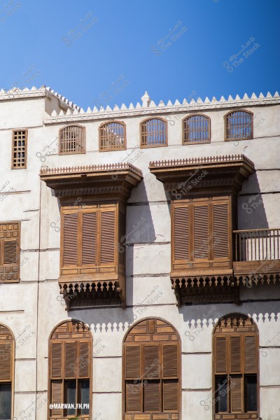 An image of a traditional building facade reflecting old architectural style in the Middle East. The building features ornate wooden windows and protruding balconies. The building is white with small domes above the windows, and a clear blue sky is in the background.