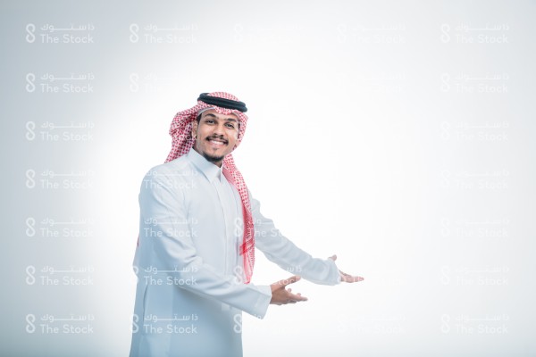 بورتريه رجل سعودي يرتدي الثوب ويقف أمام خلفية بيضاء، يتفاعل بحركات تعبيرية بيديه ويبتسم, رجل سعودي يستقبل أشخاص ويرحب بهم، صورة شخصية