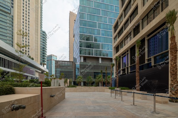 Image of a modern area adorned with palm trees, surrounded by tall buildings with glass facades and modern architecture. In the foreground is a paved walkway with palm trees lined on either side, along with a directional sign and seating arranged along the path.