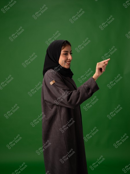 امرأة ترتدي حجاباً أسود وعباءة داكنة تشير بيدها أمام خلفية خضراء.