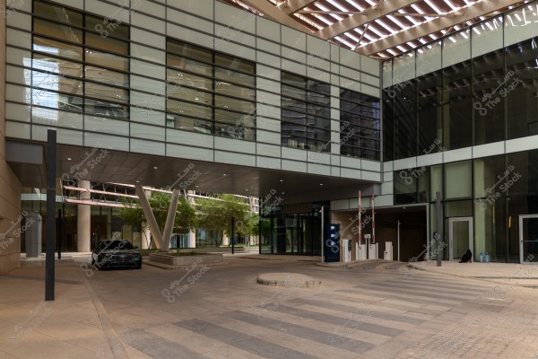 The image shows a modern courtyard of an office building, adorned with large glass facades and contemporary architectural design. A green tree is visible, and a black car is parked near a covered entrance, with entry barriers and a section of pavement marked with pedestrian lines.
