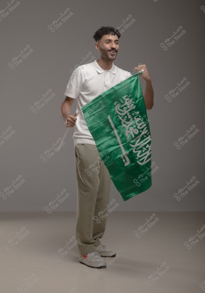 A photo of a man celebrating while holding the green flag of Saudi Arabia featuring the shahada. The man is wearing a white polo shirt, beige trousers, and sneakers. The light gray background accentuates the details, making the image appear to be taken in a studio setting.