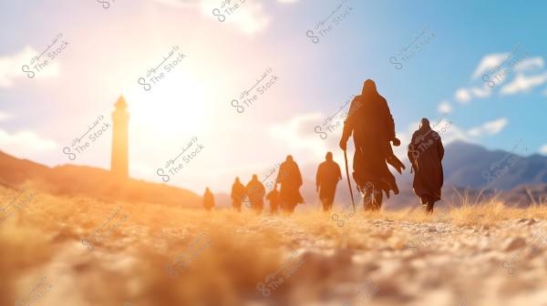 A group of people walking on a sandy path towards a tower or minaret in the distance. The sun is bright in the blue sky, casting a warm glow on the landscape. The individuals are wearing dark, loose-fitting clothing suitable for a desert or rural setting. Mountains are visible in the background.