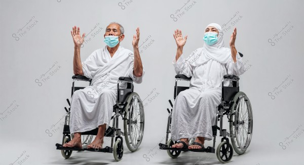 A man and a woman sitting in wheelchairs, wearing white Ihram clothing and blue face masks. They appear to be raising their hands in prayer. The background is white.