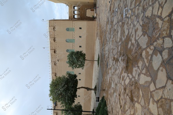 An image of a traditional mud building with green wooden windows, surrounded by a paved stone courtyard. Several green trees are scattered in the courtyard. The sky is clear and blue.