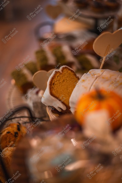 An image of a slice of cake with icing, with a blurred background featuring a variety of baked goods including a scone, next to a pumpkin-shaped decoration.