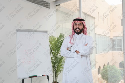 رجل سعودي يرتدي الزي السعودي التقليدي يقف مكتوف اليدين أثناء إجتماع في العمل نهارًا