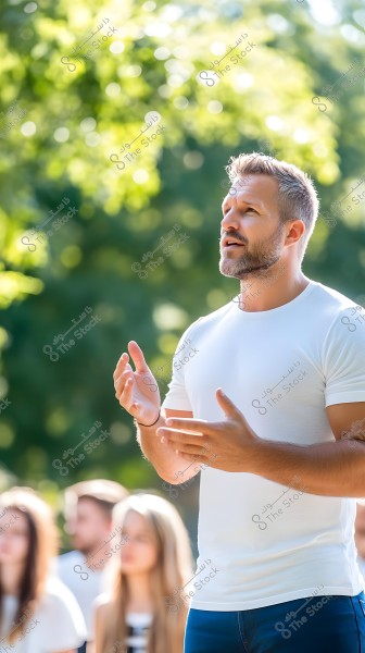 Image of a man wearing a white t-shirt standing outside in sunlight, gesturing with his hands while looking away. The background features green trees with sunlight filtering through the leaves and indistinct people in the background.