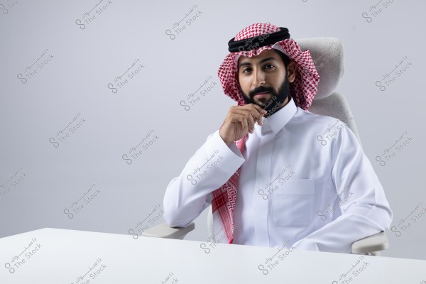 A portrait of a man sitting on a chair wearing traditional Saudi attire, including a white thobe, red-checkered ghutra, and black agal. The man appears friendly and thoughtful, with one hand on his chin, and a neutral white background behind him.