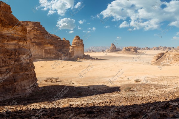 A natural landscape of a desert featuring stunning rock formations under a blue sky with scattered white clouds. The rocks are in various shades of brown surrounded by stretches of golden sand and scattered trees in the distance.