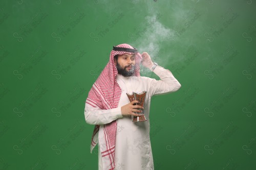 رجل سعودي يقف على خلفية خضراء يرتدي الثوب و الشماغ، يقوم بعمل حركات بيديه ، يبخر نفسه بالطيب و العود، احتفالات العيد