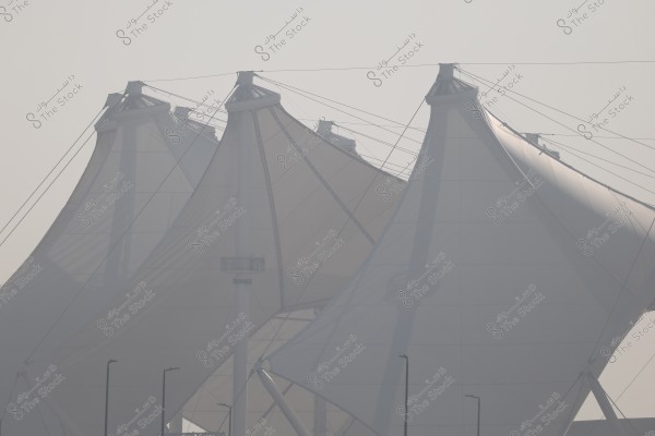 The image shows a group of large sail-like structures made of reinforced fabric, standing prominently in a foggy atmosphere. Supporting poles and cables holding the covers in place are visible. The design is characterized by its unique and modern shape.