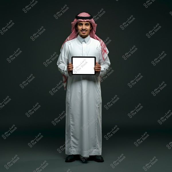 A portrait of a man standing, wearing traditional Saudi attire, including a white thobe and a red and white ghutra. He is holding a tablet with a blank white screen and is smiling against a dark background.