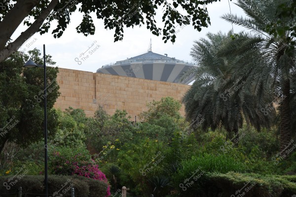 Image of a green garden featuring various trees and colorful flowers. In the background, a large water tower with a striped design is visible behind a brown stone wall. In the foreground, there are palm trees and dense plants, with a street light visible on the side of the image.