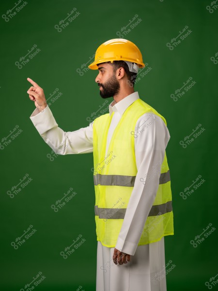 رجل يرتدي سترة أمان صفراء وخوذة واقية صفراء يشير إلى الأمام بإصبعه, يقف أمام خلفية خضراء.