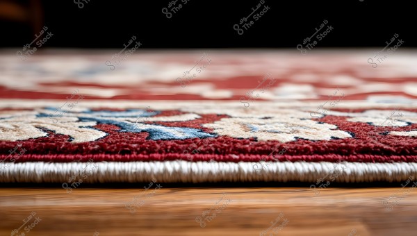 Close-up image of the edge of a carpet on a wooden floor. The carpet features a floral design with red, blue, and beige colors, with white borders on both sides. The image highlights the detailed fibers and texture of the carpet.