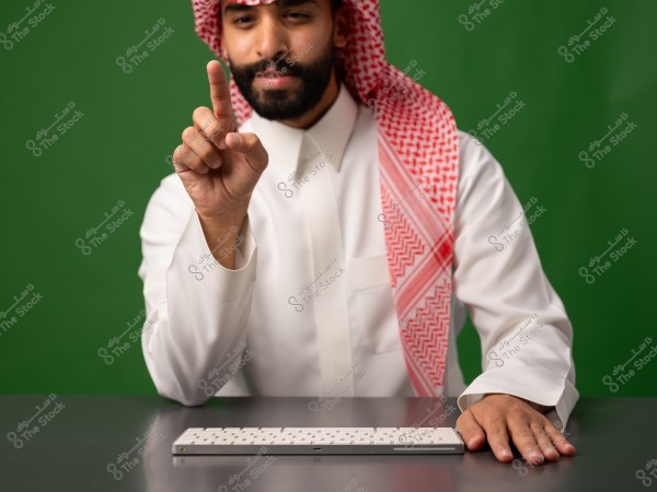 A man wearing traditional attire and keffiyeh pointing with his finger, with his other hand on a keyboard on a table, on a green background.