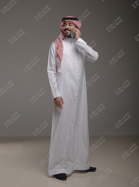 Image of a man wearing a white Saudi thobe with a red checkered shemagh and a black agal, standing against a gray background and talking on the phone. He appears smiling and relaxed.