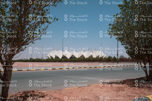 The stadium of King Fahd International Stadium in Riyadh by day, and in front of it an iron fence and a lot of trees