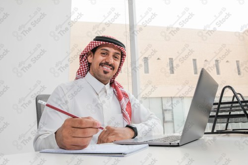 رجل سعودي يرتدي الزي السعودي التقليدي يعمل في مكتب بواجهة زجاجية ويستخدم قلم رصاص لتدوين الملاحظات