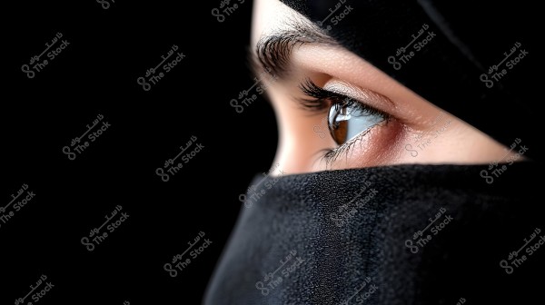 Portrait image showing the eye of a person wearing a hijab covering the head and neck. The focus is on the eye with long eyelashes, and the black background highlights the facial features beautifully. The traditional hijab reveals a small part of the face.