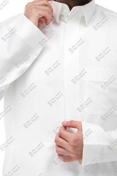 Image of a man wearing a white thobe, showing the upper body and sleeves. The thobe appears modern and elegant with a traditional collar and long sleeves. The man is fastening the collar button with his right hand.