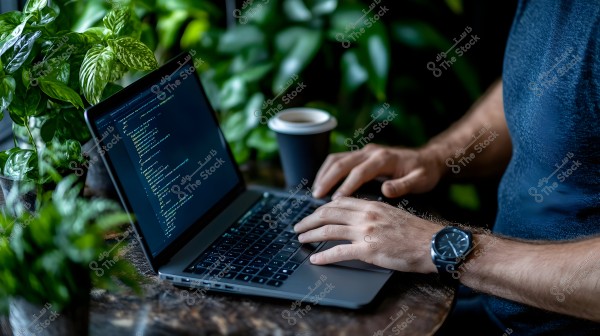 The image shows a person working on a laptop with a screen displaying code. The person appears to be seated at a wooden table in an environment surrounded by several green plants. They are wearing a black watch and a dark blue shirt, with a coffee cup beside them.