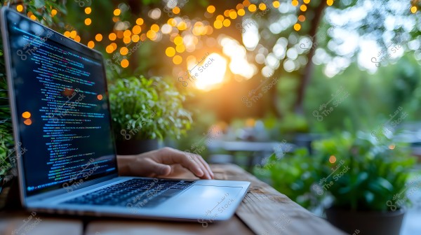 An open laptop on a wooden table in an outdoor setting. The laptop screen displays colorful coding text. The background shows a bokeh effect with warm lights and green trees under sunlight.