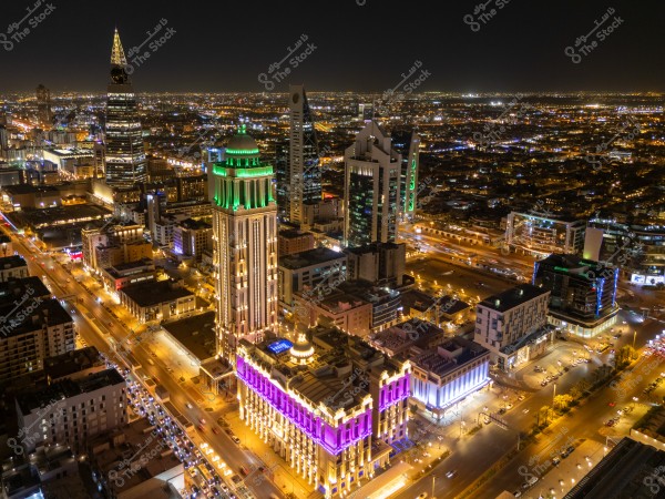 The image shows an aerial night view of Riyadh, Saudi Arabia. Prominent in the image are towers illuminated in multiple colors such as green and purple, surrounded by lit streets and urban buildings. The horizon extends far with city lights illuminating the sky.