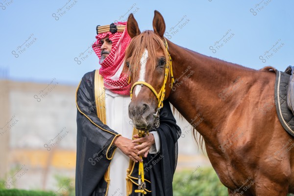 A portrait of a man wearing traditional Saudi attire standing next to a brown horse. The man is dressed in a white robe with a red headscarf and black agal, as well as a black bisht with golden embroidery. The horse is adorned with a yellow bridle around its head. The background features a clear blue sky.