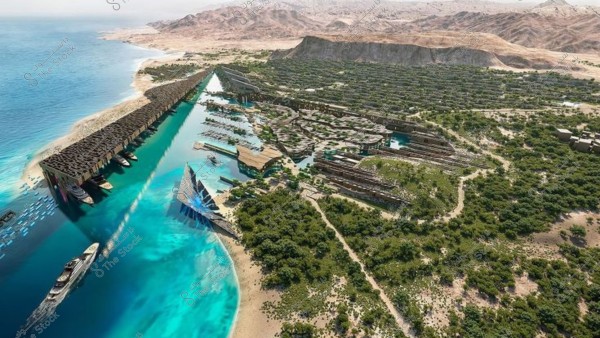 صورة جوية لمشروع معماري حديث يقع على شاطئ بحر أزرق. يظهر في الصورة مبانٍ بتصاميم مبتكرة تمتد بمحاذاة الساحل, تتخللها قنوات مائية تتصل بالبحر. المنطقة محاطة بالأشجار والمسطحات الخضراء, وتظهر خلفية الصورة جبال صخرية بنية.