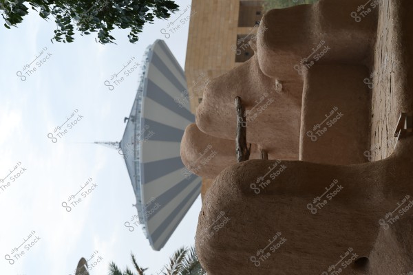 An image of a traditional mud-brick building, with a circular roof structure in the background featuring a striped design. The building appears to be part of a heritage site surrounded by palm trees. The sky is slightly cloudy, creating a serene atmosphere in the image.