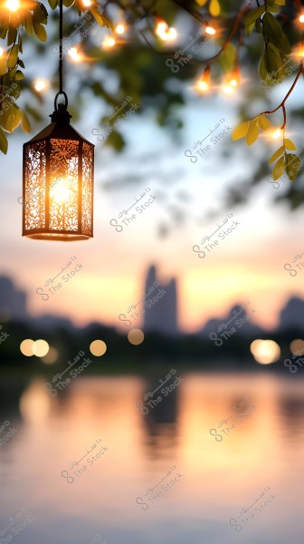 An illuminated lantern with an ornate design hanging from a tree branch, surrounded by small twinkling lights. The background features a sunset with horizontal reflections and blurred silhouettes of city buildings, creating a calm and beautiful atmosphere.